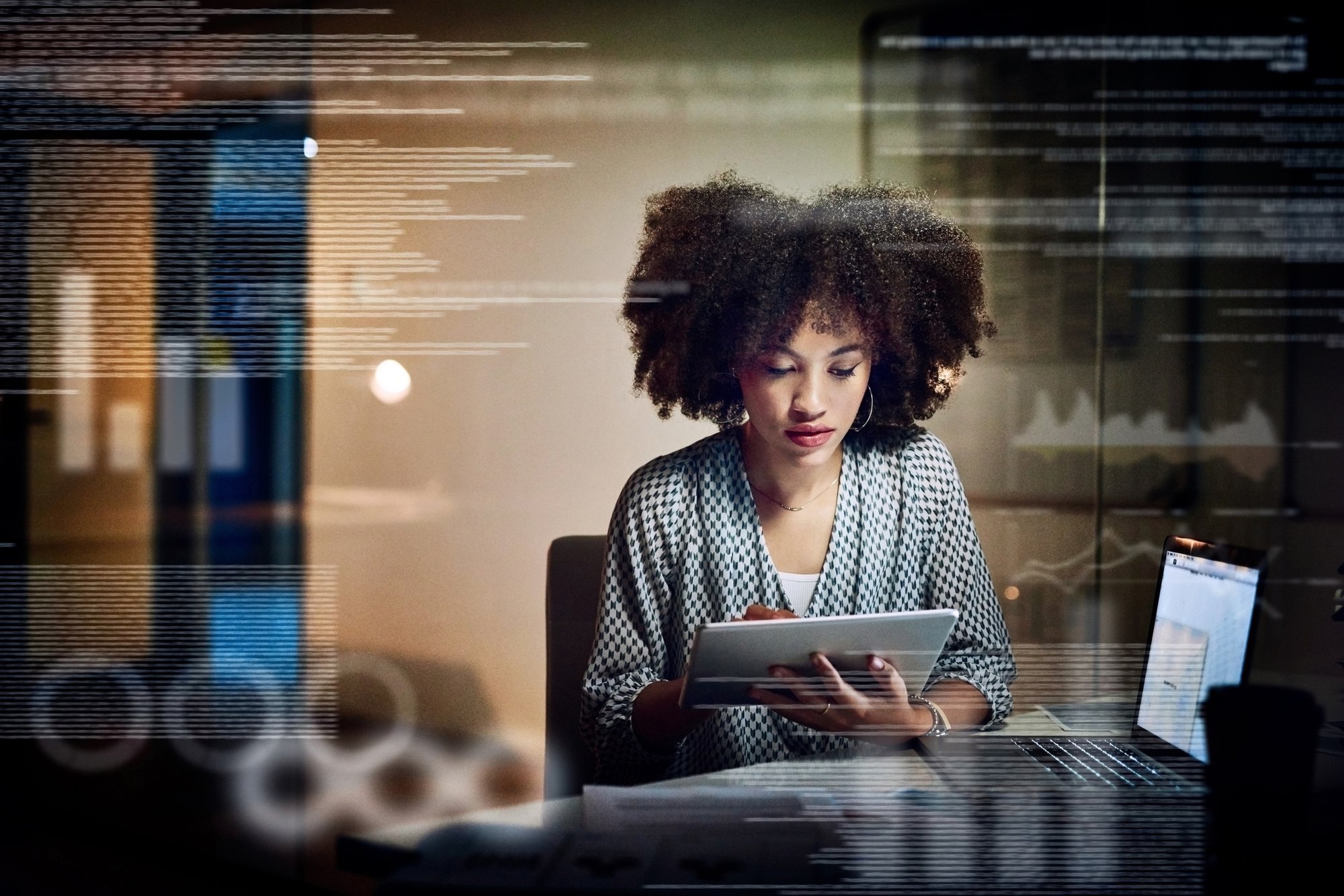 Woman holding a tablet at a desk with data and graphs juxtaposed on top.